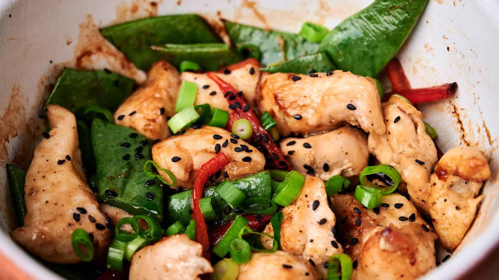 A close-up of a stir-fry dish featuring pieces of chicken, snap peas, red bell peppers, scallions, and black sesame seeds.