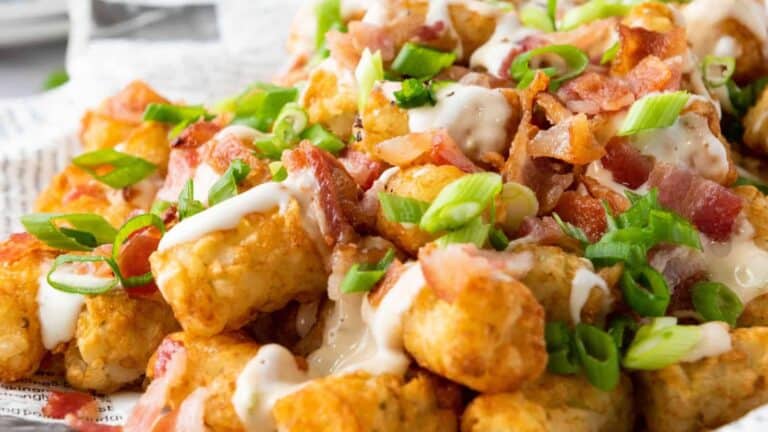 Close-up of crispy tater tots topped with melted cheese, crispy bacon pieces, sliced green onions, and drizzled with a creamy white sauce.