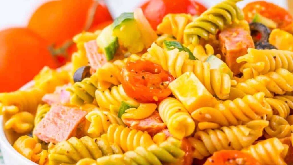 Close-up of a colorful pasta salad with rotini, cherry tomatoes, chopped vegetables, cubes of ham, black olives, and garnished with herbs. Tomatoes are visible in the background.