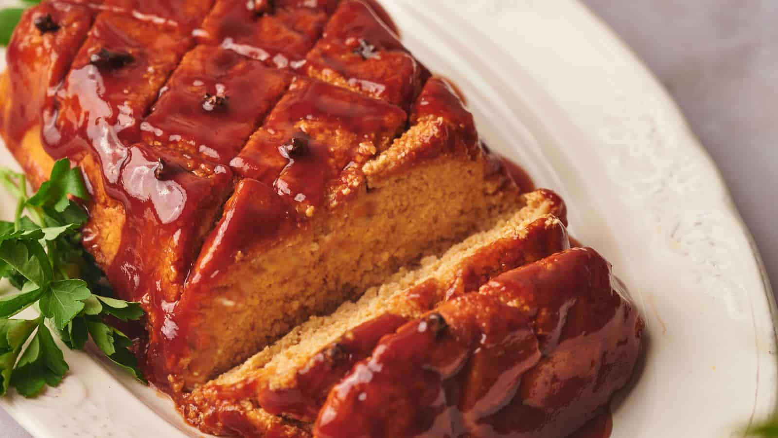 A glazed meatloaf with a crosshatch pattern sits on a white platter, garnished with parsley. Two slices are cut from one end.