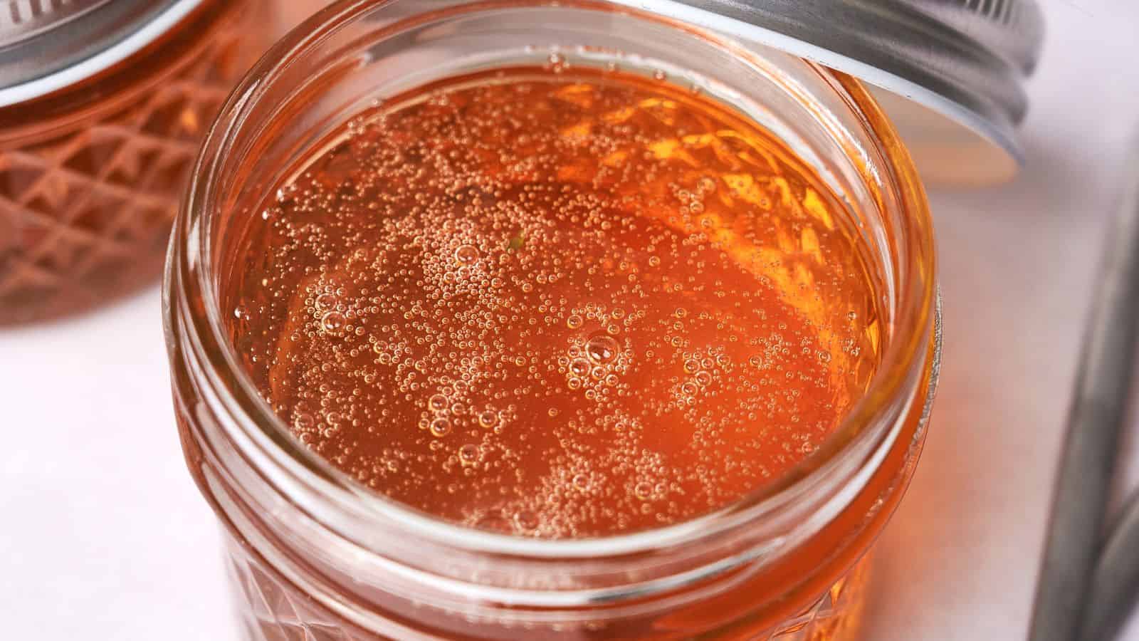 Close-up view of a jar filled with golden, translucent vegan honey and numerous tiny air bubbles, set on a tabletop, with a wire basket visible in the background.