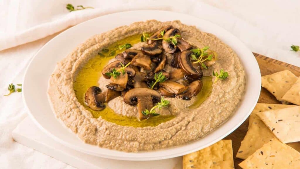 A bowl of hummus topped with sautéed mushrooms and herbs, served with crackers on a white tablecloth.