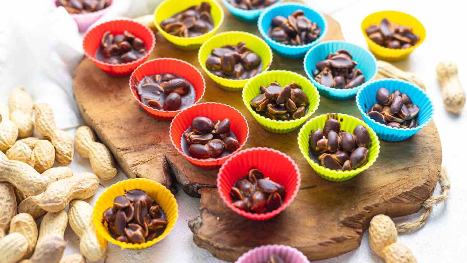 Colorful mini cupcake liners filled with chocolate and peanuts on a wooden board, surrounded by raw peanuts.