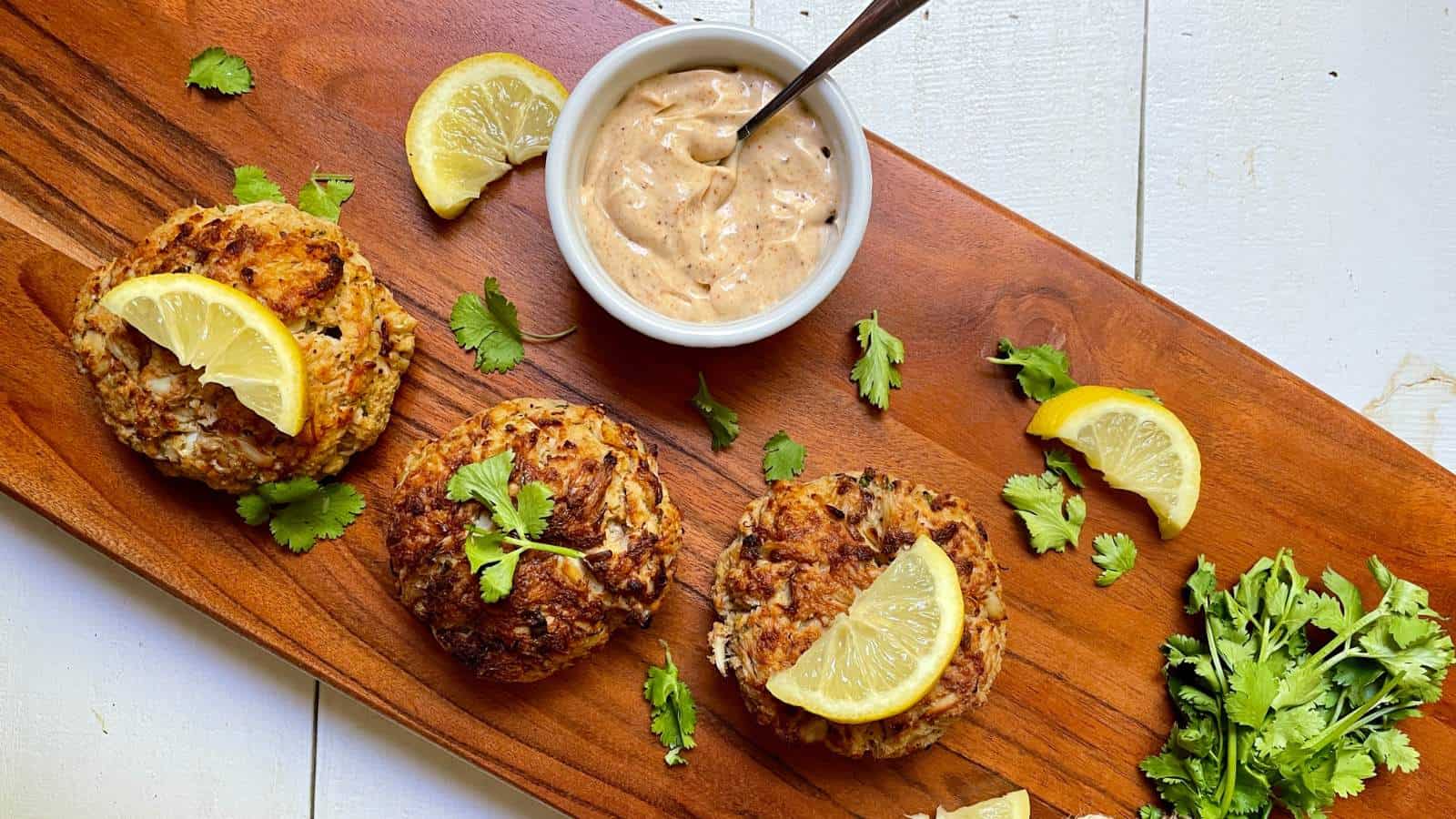 Three crab cakes garnished with lemon wedges and parsley are arranged on a wooden board, accompanied by a small bowl of dipping sauce with a spoon.