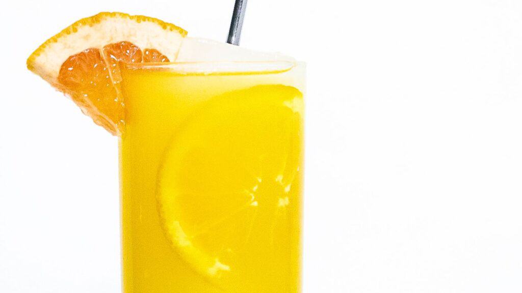 A close-up image of a glass of orange juice with a slice of orange on the rim and a grapefruit wedge on top, against a white background.