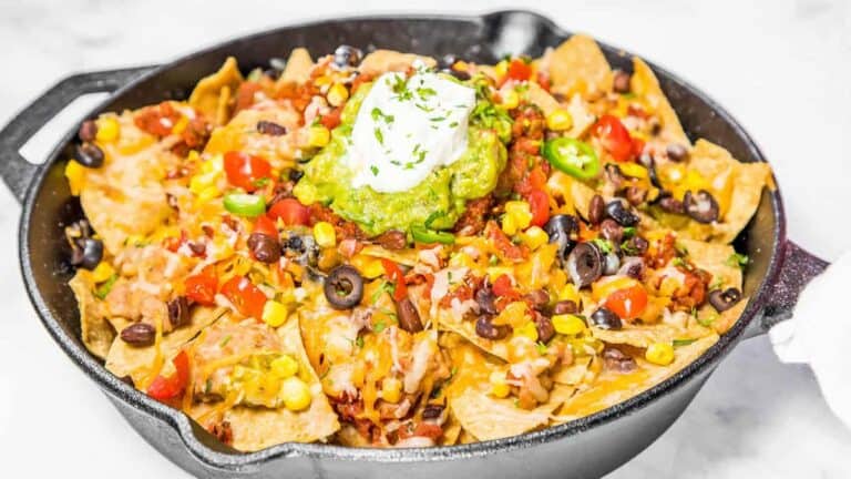 A skillet of nachos topped with cheese, jalapeños, olives, corn, diced tomatoes, guacamole, and sour cream.