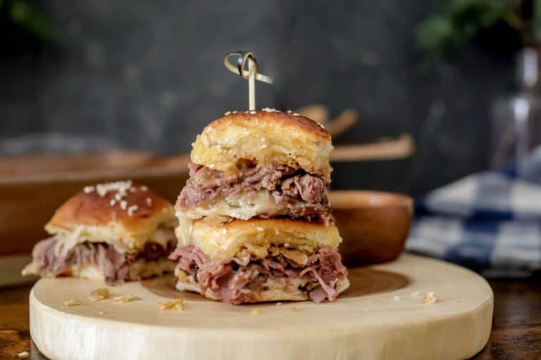 Two small roast beef sliders are stacked on a wooden board, secured with a toothpick. Another slider is in the background. The sliders have sesame seed buns and appear to be served in a rustic setting.
