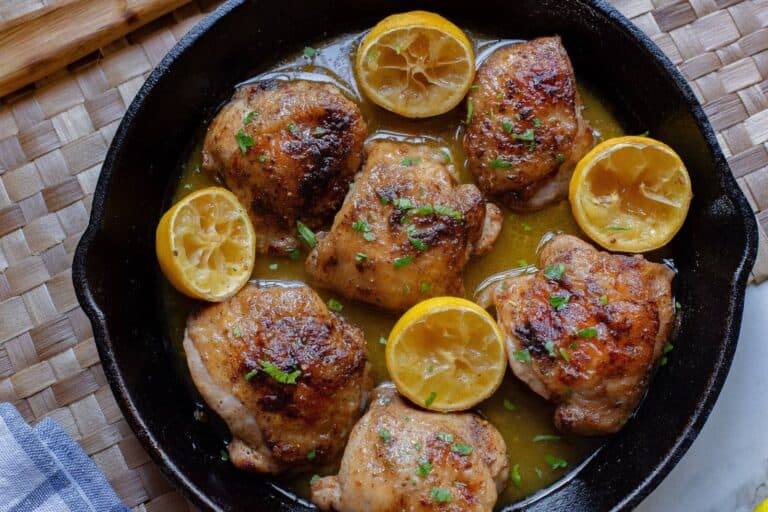 A cast iron skillet with six cooked chicken thighs garnished with parsley and halved lemons.
