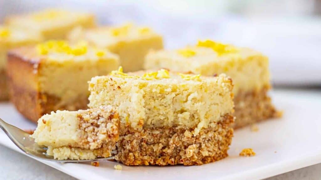 A slice of lemon crumb bars on a white plate, featuring a thick crumbly base and a zesty lemon topping with visible lemon zest garnish.