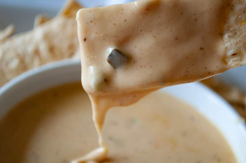 A chip, adored by all who loves cheese, is dipped in creamy cheese sauce with visible spices and green pepper pieces, held above a bowl filled with the same flavorful sauce, with other chips in the background.