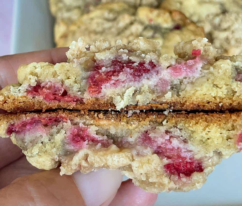 Hand holding a freshly baked cookie, split in half, showing its soft interior with vibrant pink raspberry filling.