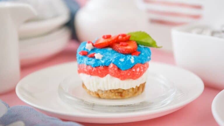 A small dessert in patriotic colors with a layered base of creamy white, red, and blue topped with strawberry slices and mint, served on a white plate.