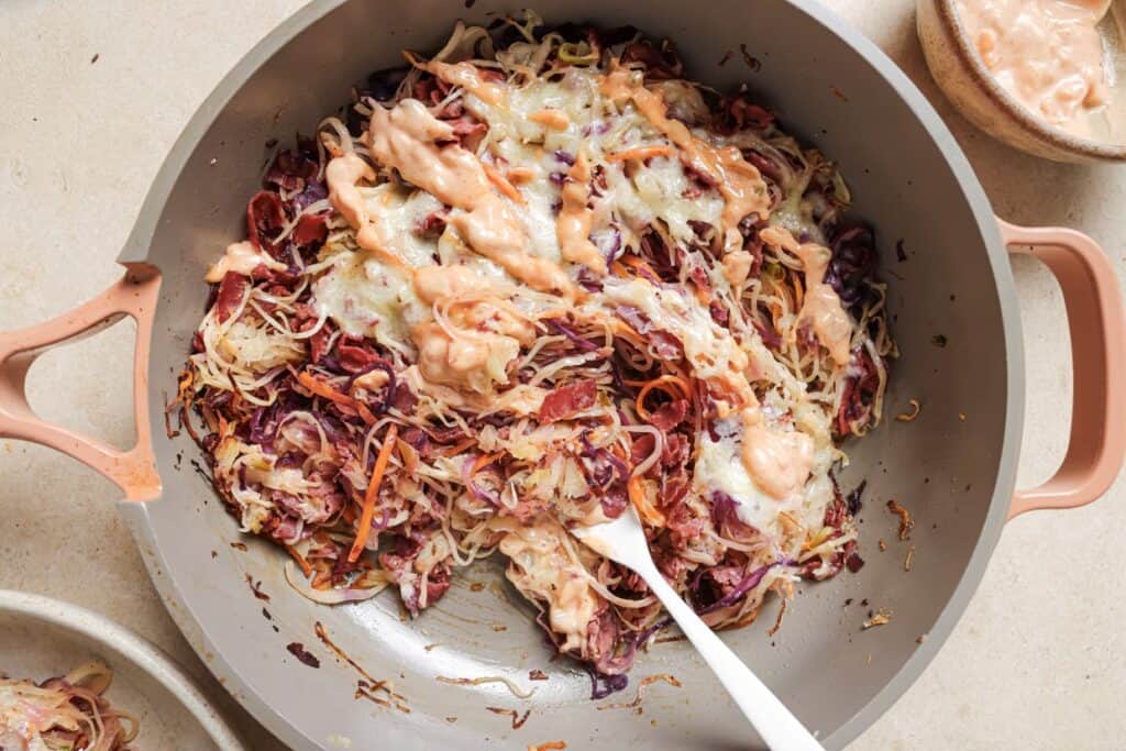 A skillet with sautéed red and white cabbage, topped with melted cheese and a drizzle of creamy sauce, stirred by a white spatula.