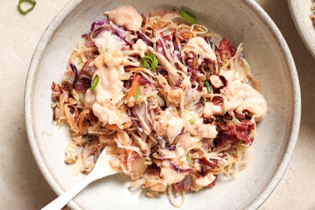 A bowl of coleslaw with creamy dressing, garnished with green onions, served on a light beige surface.