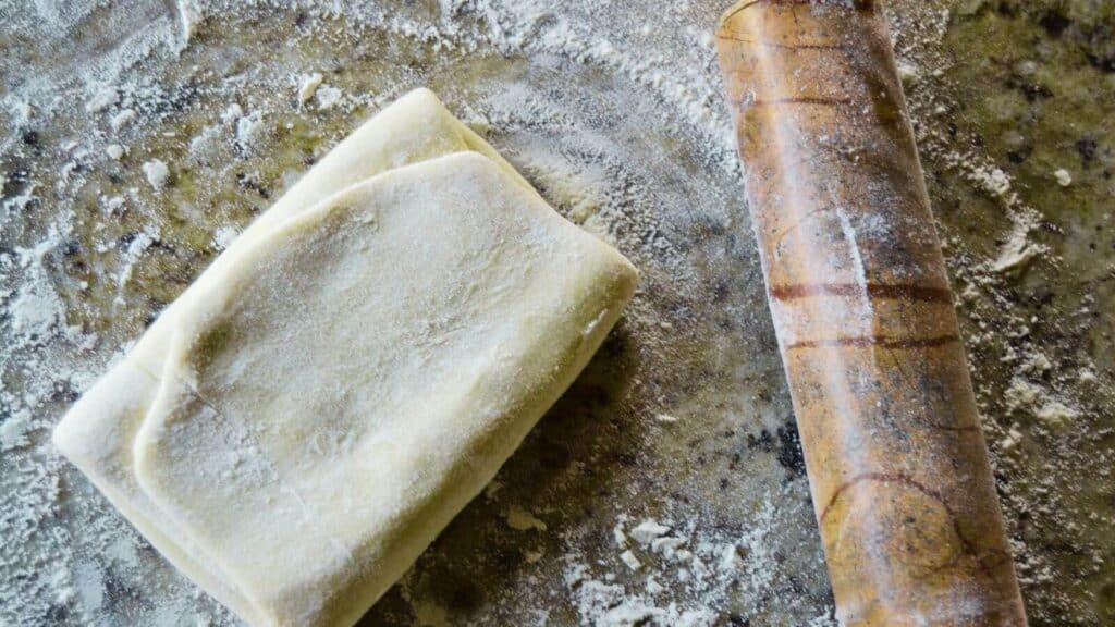 A piece of folded puff pastry dough rests on a floured surface next to a wooden rolling pin.