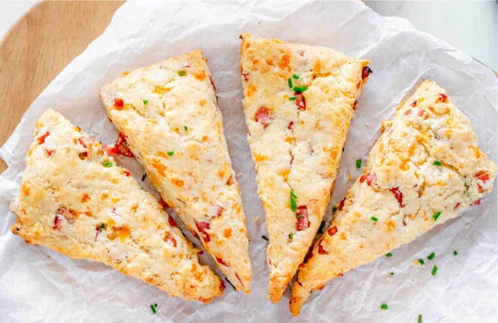 Four triangular scones with visible chopped red and green ingredients are arranged on crinkled parchment paper, on a wooden board.