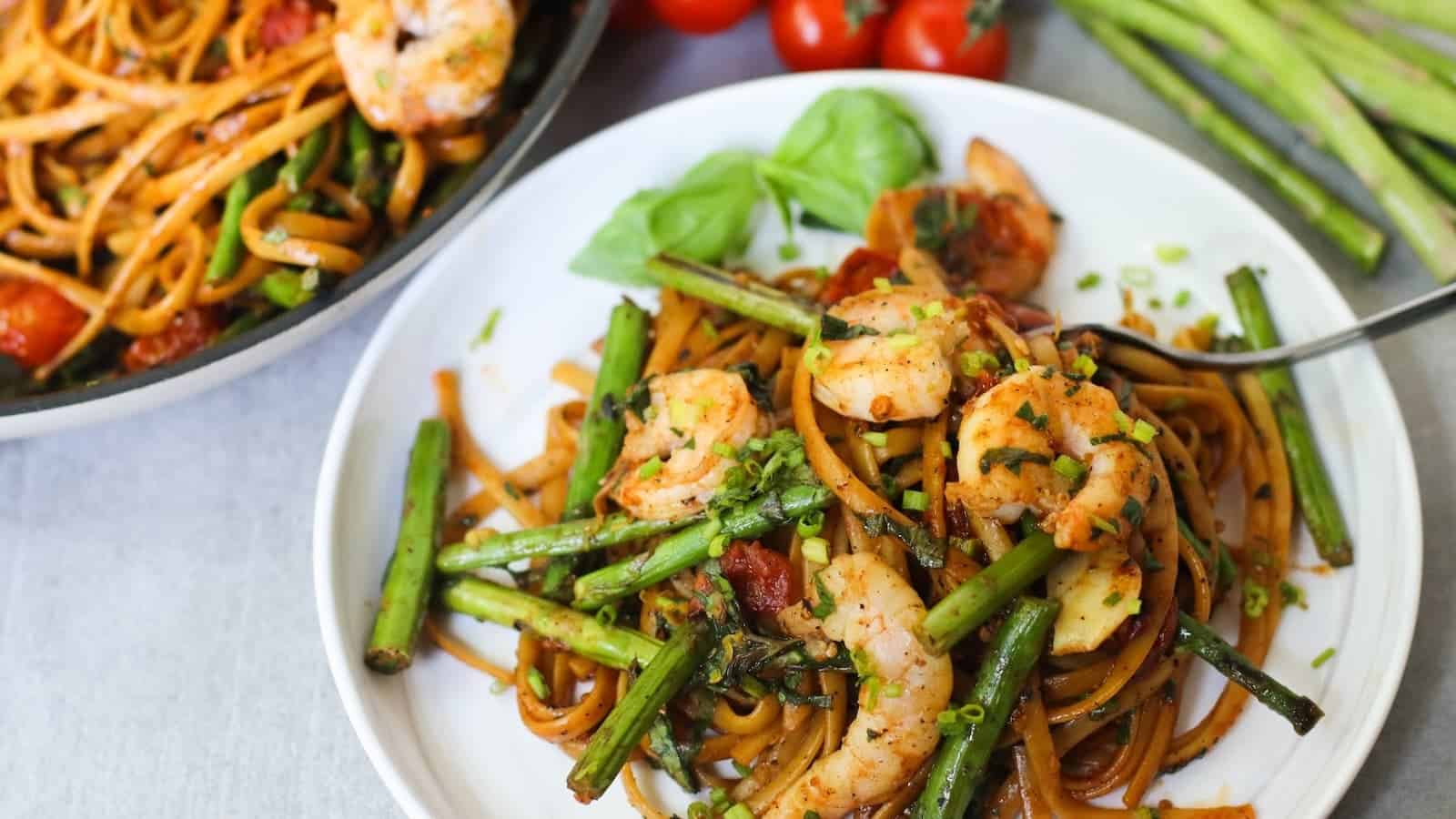 A plate of pasta with shrimp, asparagus, tomatoes, and a garnish of fresh herbs. A skillet with more pasta and additional tomatoes and asparagus can be seen in the background.