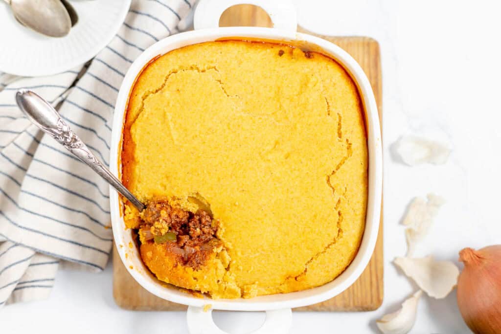 A casserole dish with golden cornbread topping and a spoon scooping out a portion, revealing a ground meat filling beneath. The dish is placed on a wooden board with a striped cloth nearby.