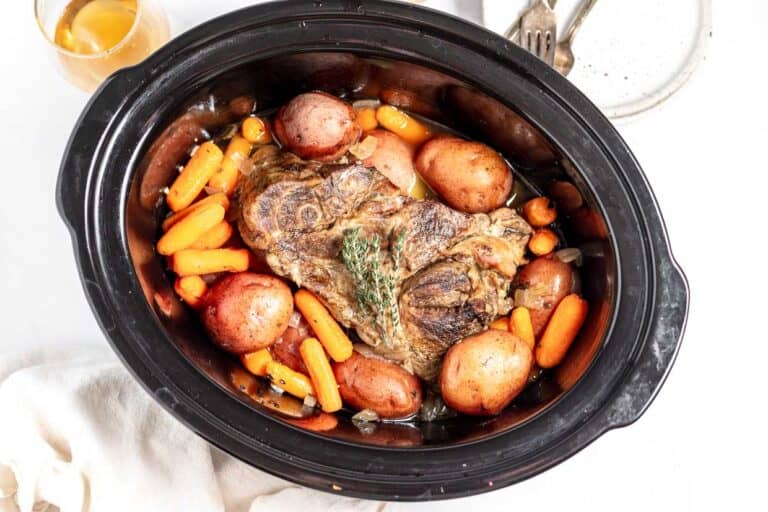 Top view of a slow cooker with beef roast, carrots, and potatoes. a small bowl of gravy and utensils are on the side.