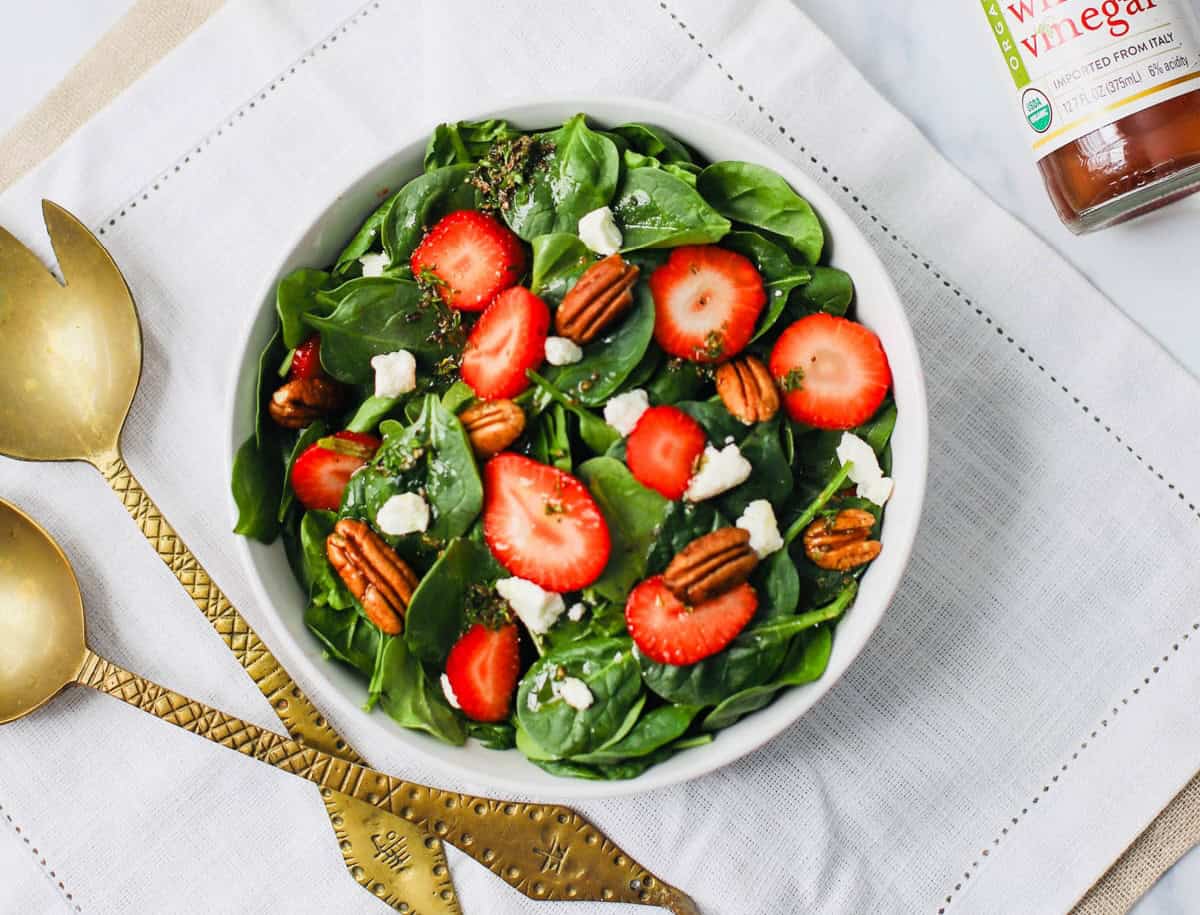 Spinach with strawberries, goat cheese, pecans, and red wine vinaigrette in a white bowl on a white napkin with bottle of red wine vinegar and bronze serving set.