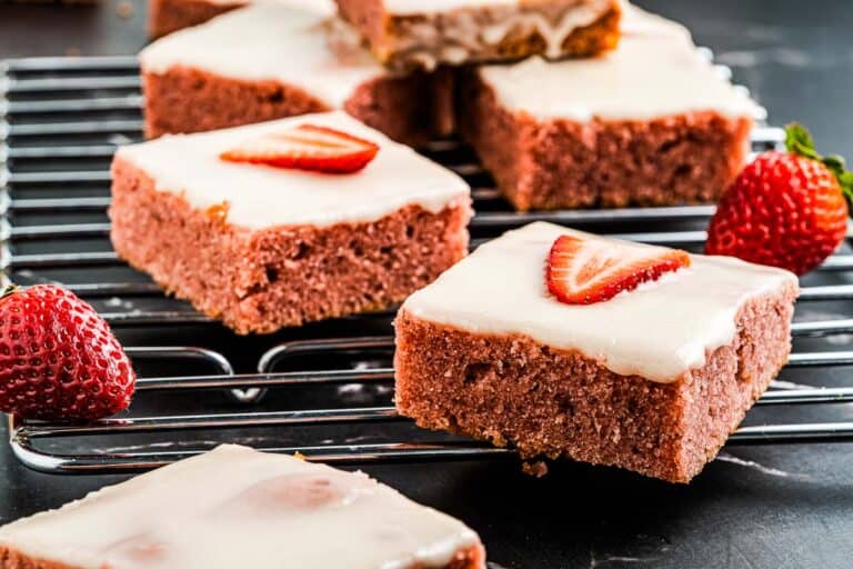 Slices of strawberry-topped frosted brownies on a cooling rack, with fresh strawberries nearby.