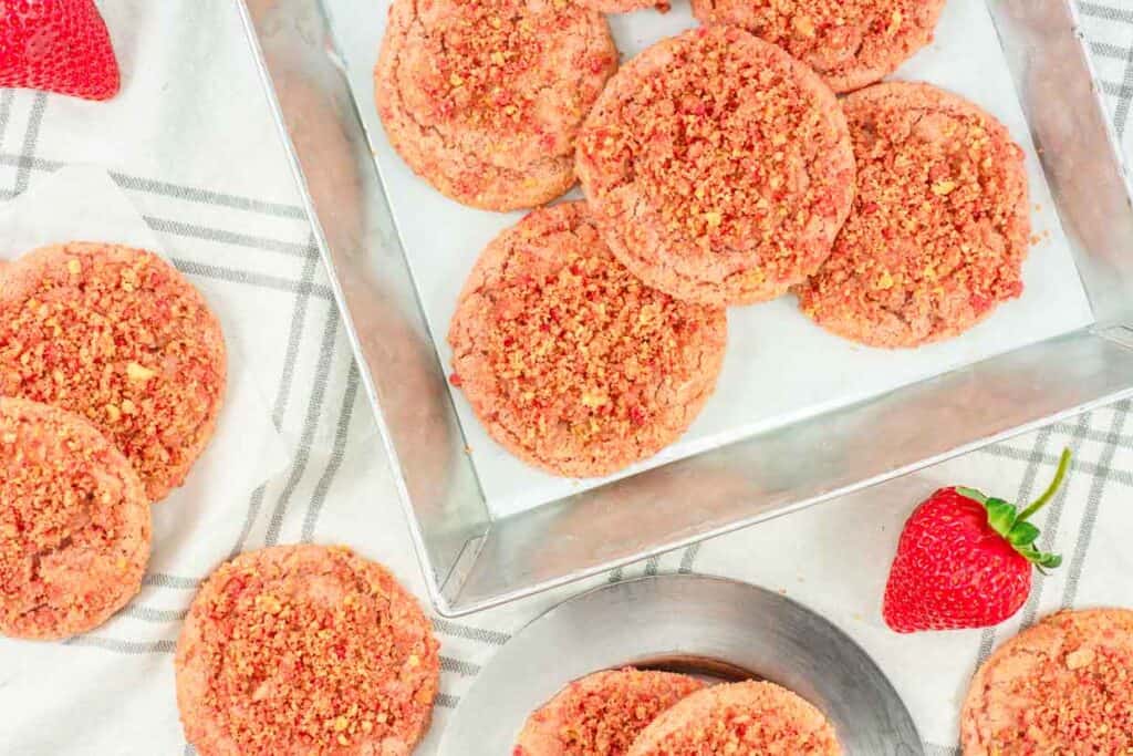 A tray of freshly baked strawberry crumble cookies next to fresh strawberries and a metal scoop on a light fabric.