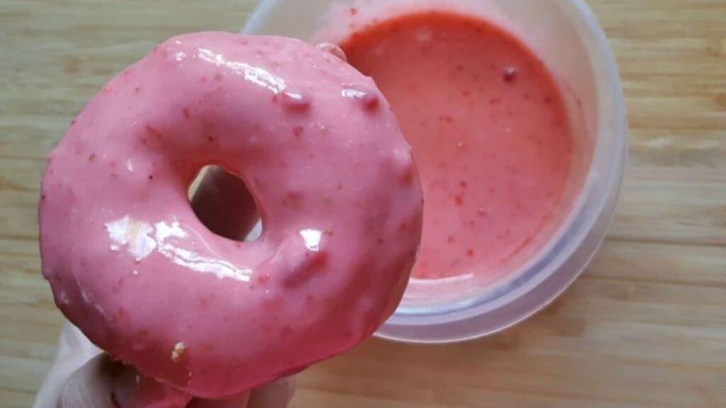 A pink frosted donut held above a small container of matching pink dipping sauce, all placed on a wooden surface.