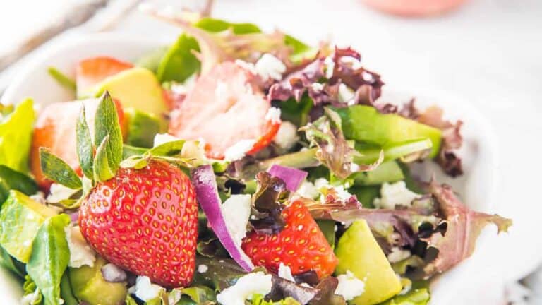 A fresh mixed salad with strawberries, avocado slices, and feta cheese on a white plate, with a light background.