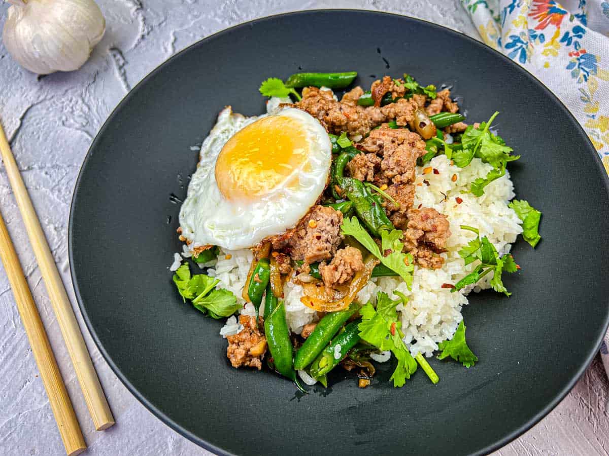 A plate of Thai pork & Green beans with rice, topped with a fried egg, served with chopsticks on the side.