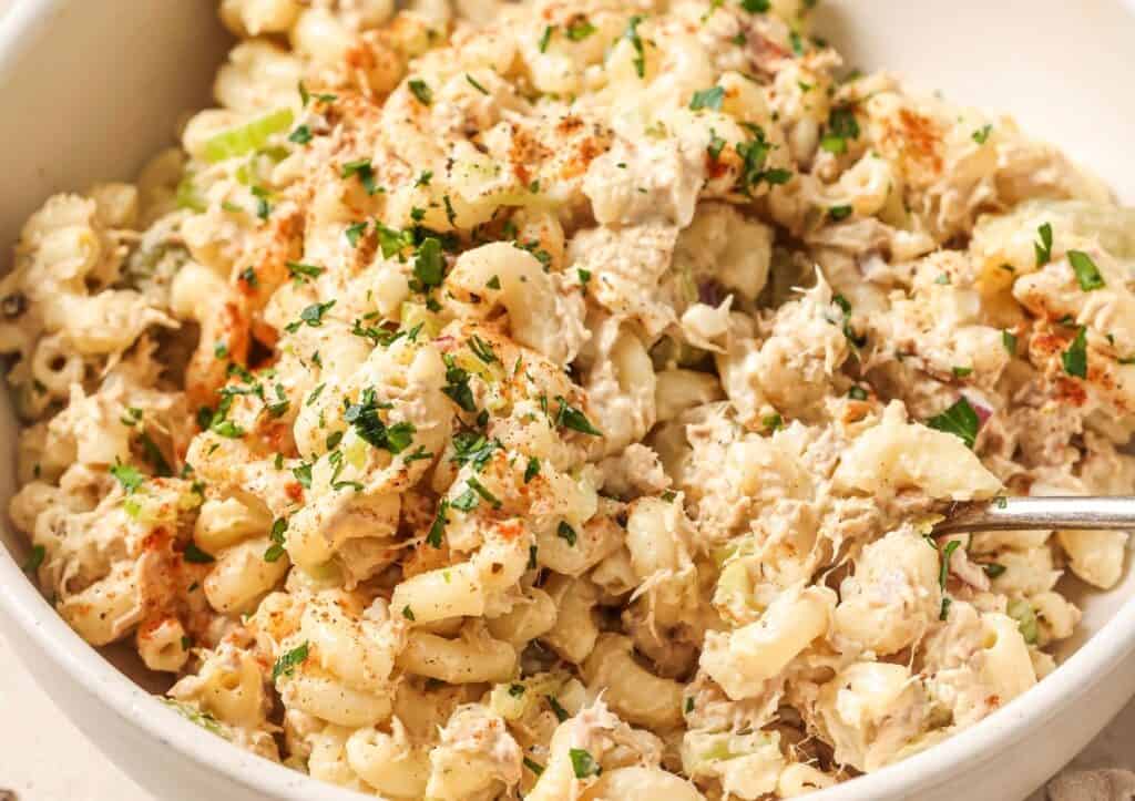 Close-up image of a bowl of tuna macaroni salad sprinkled with paprika and garnished with chopped parsley.