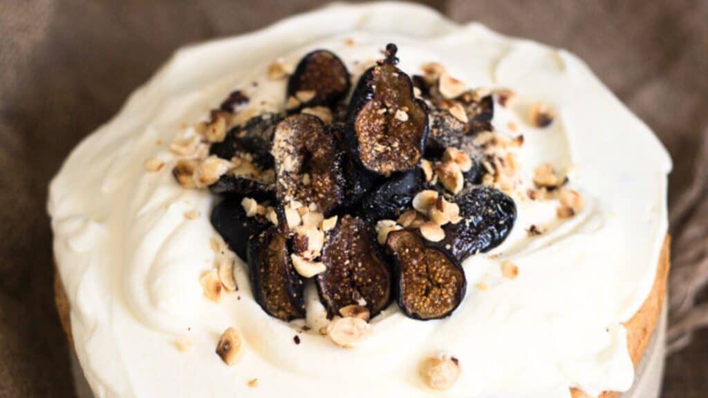 A close-up of a cake topped with white frosting, sliced figs, and crushed nuts.
