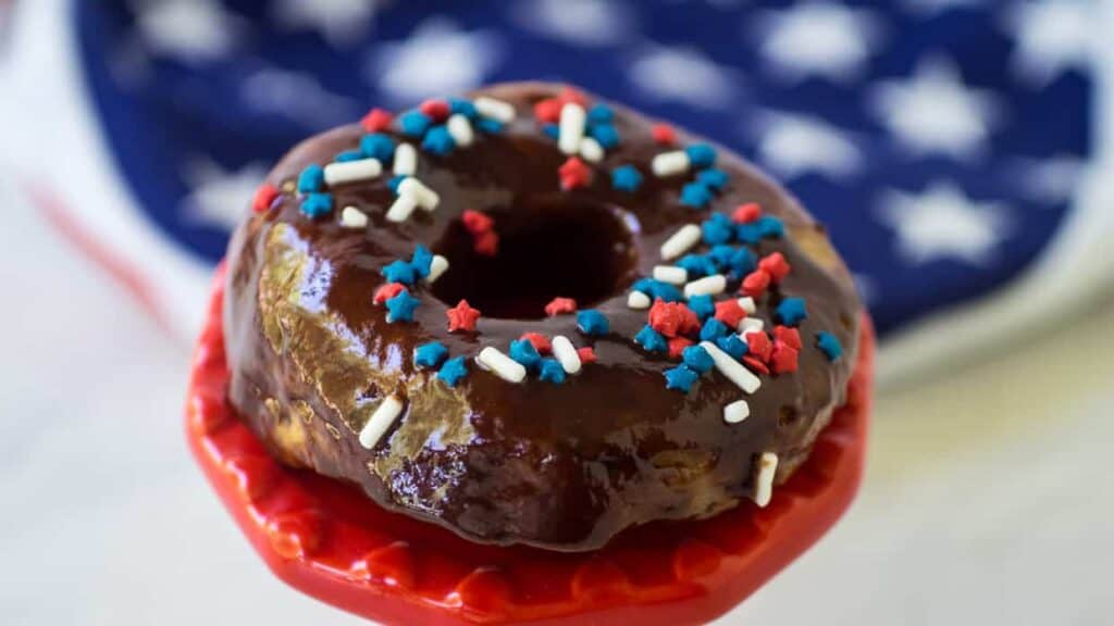 A chocolate-glazed donut with red, white, and blue sprinkles sits on a red pedestal, with a blue and white star-patterned background.