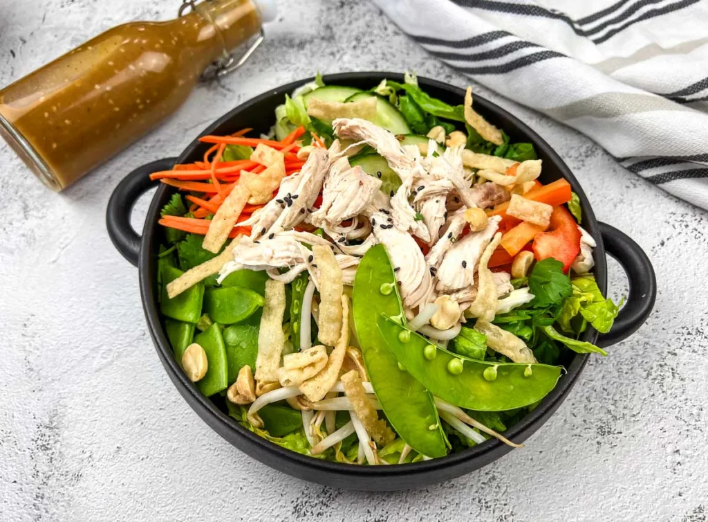 A bowl of Chinese chicken salad with shredded chicken, snap peas, carrots, and peppers, accompanied by a bottle of dressing on the side.