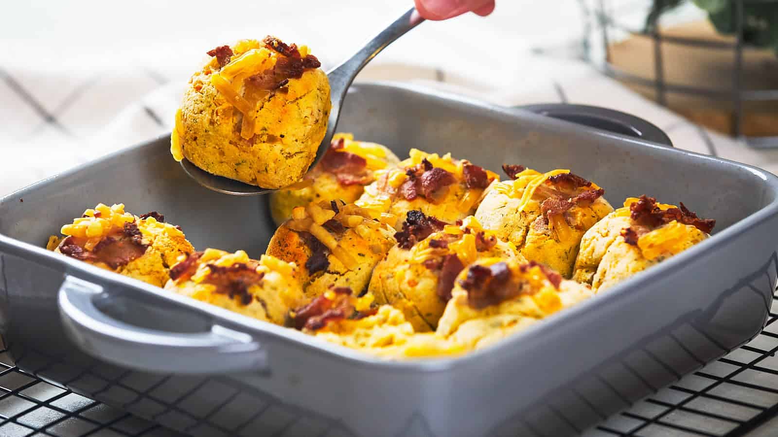 A hand scooping a serving of baked cheesy bread rolls topped with bacon from a rectangular dish.