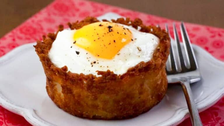 A cooked egg sits inside a crispy hash brown nest on a white plate. A metal fork rests beside the plate on a pink patterned surface.