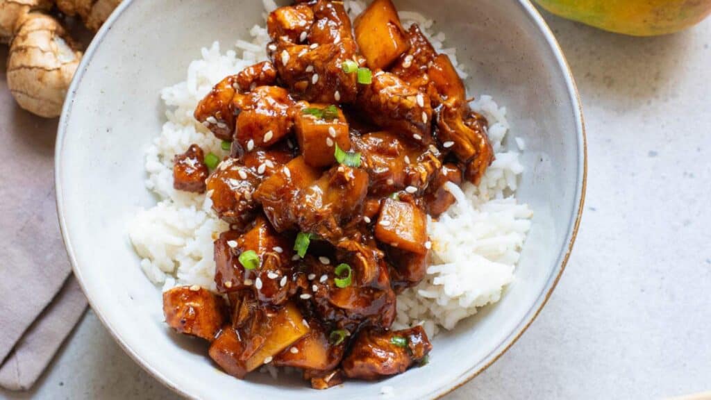 A bowl of sticky bourbon chicken in a white bowl over rice, garnished with sesame seeds.