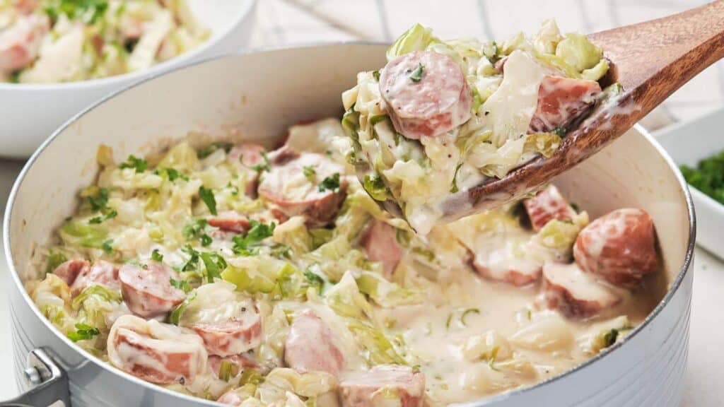 A wooden spoon lifting creamy cabbage and sausage stew from a white pot, garnished with parsley.