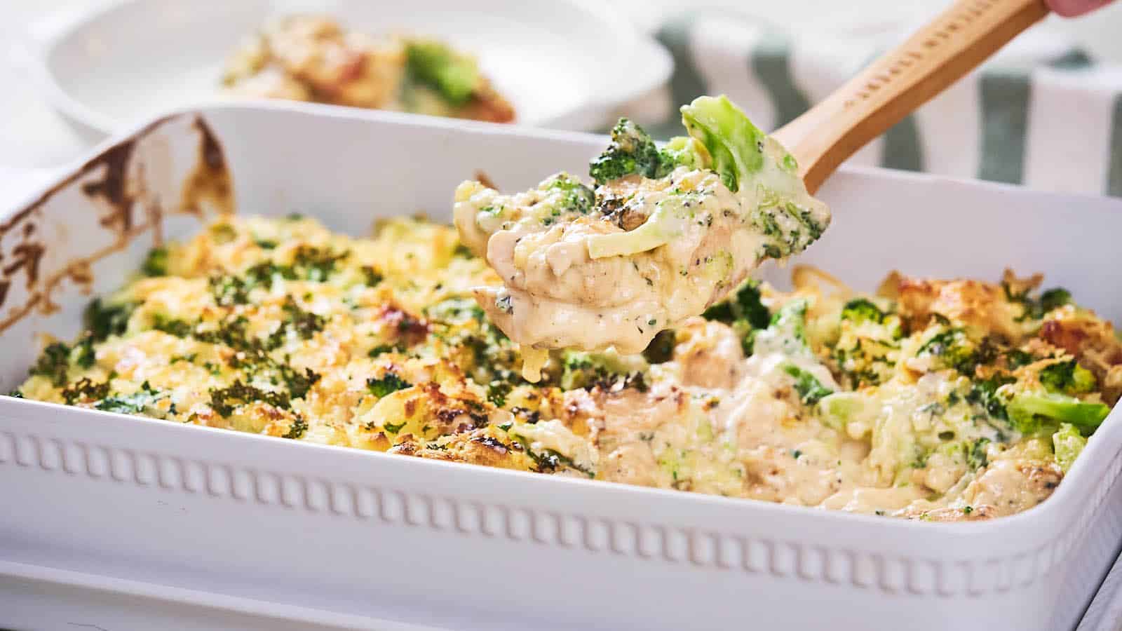 A serving utensil lifts a portion of broccoli and cheese casserole from a white baking dish.