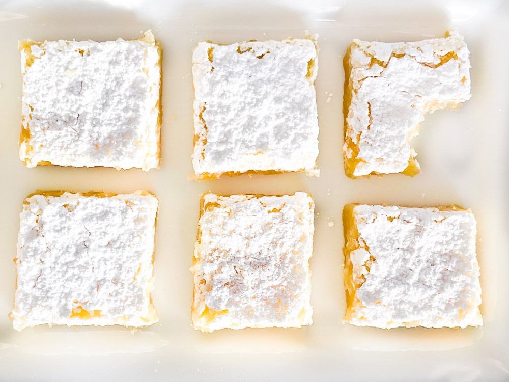 Six square lemon bars with powdered sugar topping, arranged in a white ceramic dish, viewed from above.