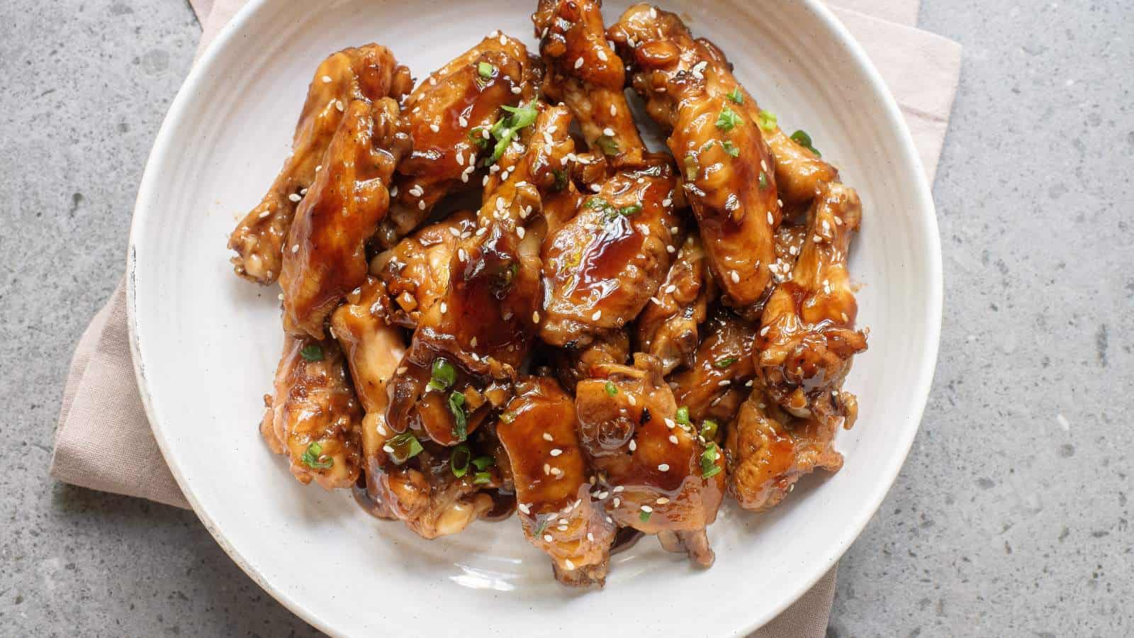 A white plate with glazed chicken wings garnished with chopped green onions and sesame seeds, placed on a gray surface.