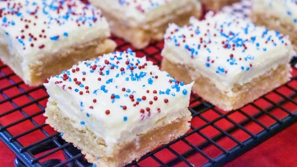 Close-up of frosted cake squares with white icing and red and blue sprinkles on a cooling rack.
