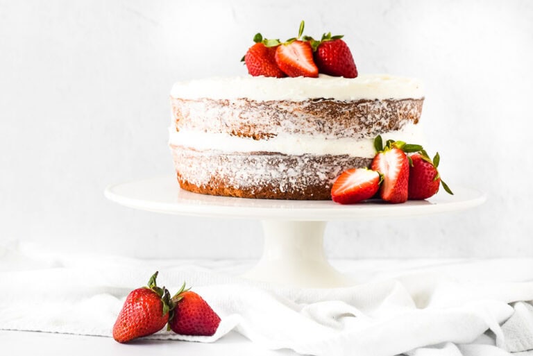 A two-layer naked cake with white frosting, topped with fresh strawberries, displayed on a white cake stand. Additional strawberries are placed on a white cloth below the stand.