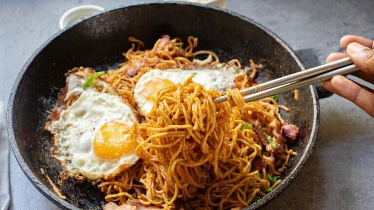 A pair of chopsticks hold a portion of cooked noodles from a skillet filled with noodles, two fried eggs, and vegetables.