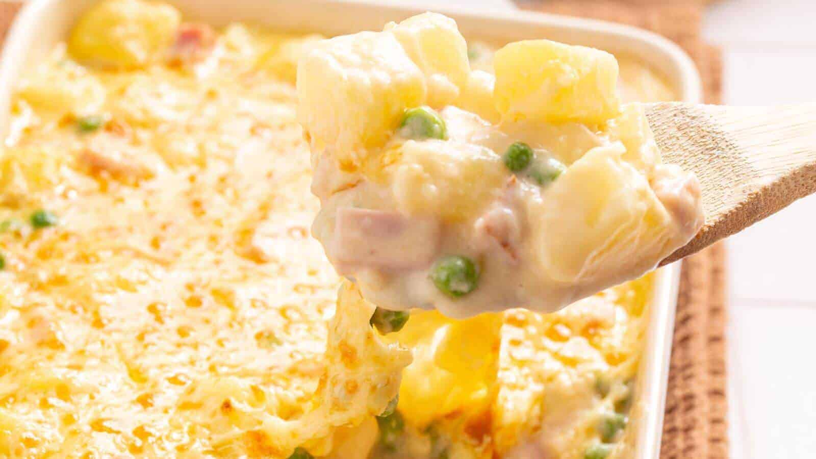 A close-up image of a spoonful of creamy ham and potato casserole with peas and cheese, lifted from a baking dish.