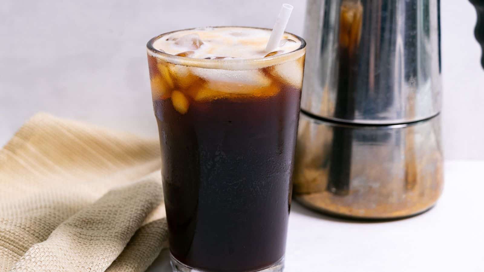 A glass of iced coffee with a straw, next to a coffee press and a beige napkin, on a light background.