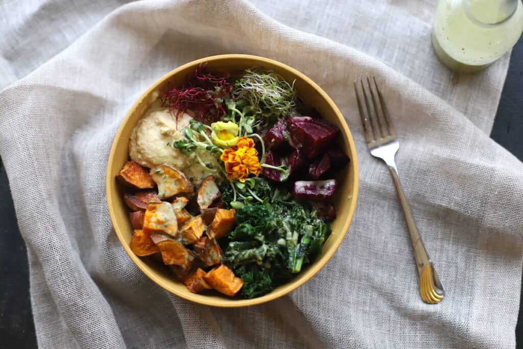 A bowl of mixed vegetables including roasted sweet potatoes, kale, beets, microgreens, and hummus, garnished with an edible flower, placed next to a fork and a small bottle on a cloth surface.