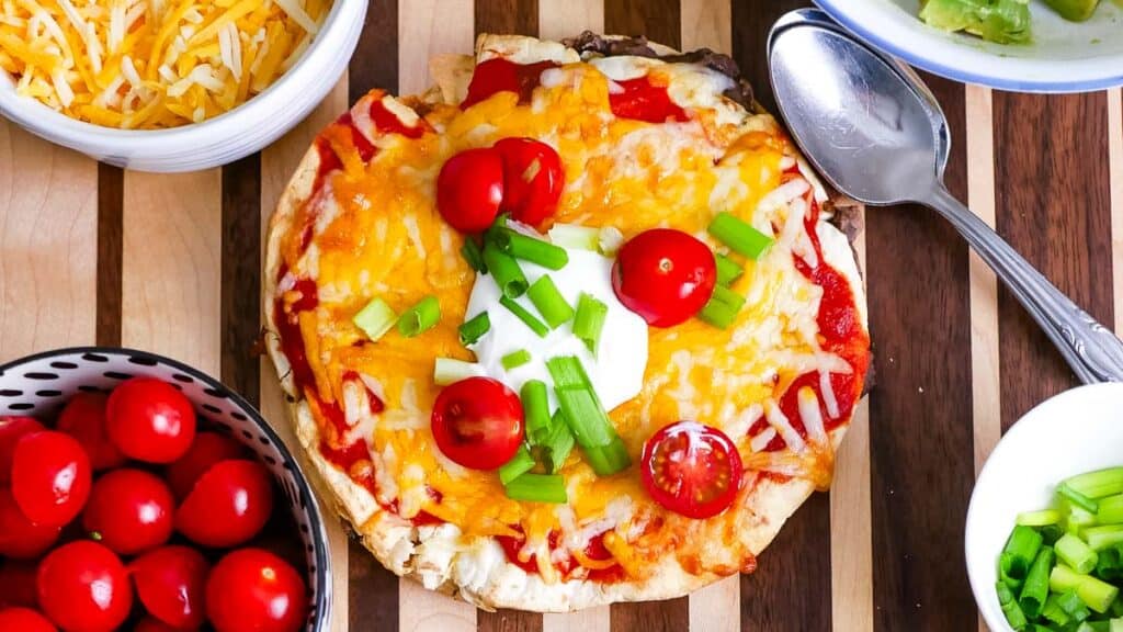 A personal pizza topped with melted cheese, cherry tomatoes, green onions, and a dollop of sour cream is on a wooden board alongside bowls of shredded cheese, cherry tomatoes, guacamole, and green onions.