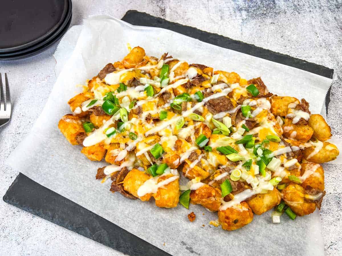 Philly Cheesesteak Loaded Tots on parchment paper, topped with Alfredo cheese, green onions, and a drizzle of white sauce, placed on a dark rectangular slate plate.