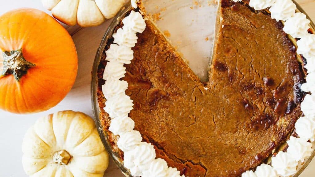 Pumpkin pie with one slice missing, decorated with whipped cream on a white plate, accompanied by whole pumpkins and garlic bulbs on a white surface.