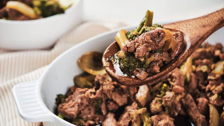 A wooden spoon scooping beef and broccoli from a white ceramic dish.